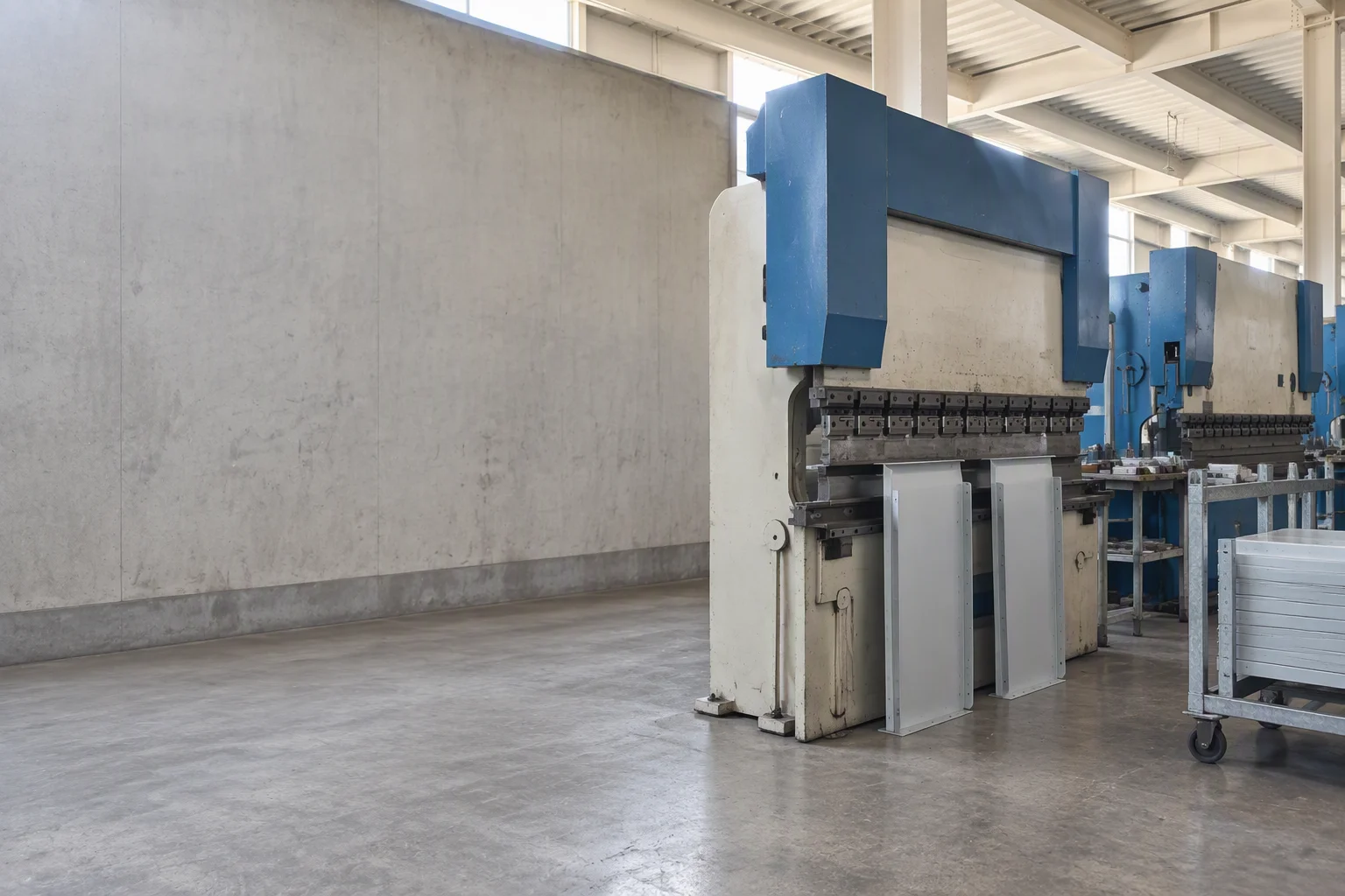 Sheet-metal workshop: two formed cabinet side panels standing on edge against the machine base, the press brake above them caught mid-stroke, a galvanised panel trolley at the right and the rest of the machine row receding.